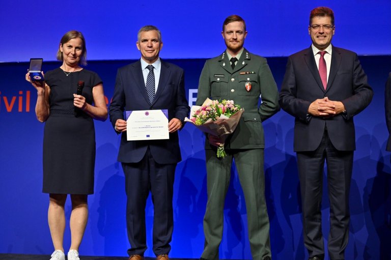 En kvinne og tre menn på en scene. Kvinnen i kjole holder en medalje, én av mennene holder frem et diplom og én mann i militæruniform holder en blomsterbukett.
